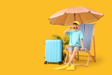 Çantalı, güvertedeki sandalyede oturan ve sarı arka planda şemsiyesi olan sevimli yüzgeçli bir çocuk. Seyahat kavramı