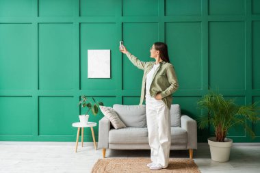 Evdeki yeşil duvarın yanında klimayı açan genç bir kadın.