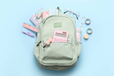 Mavi arka planda farklı kırtasiye malzemeleri ve gözlüklerle yeşil okul sırt çantası.