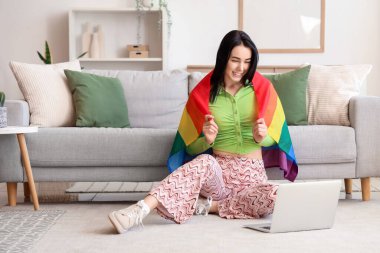 LGBT bayrağı ve modern dizüstü bilgisayarı olan güzel bir kadın oturma odasında halının üzerinde oturuyor.