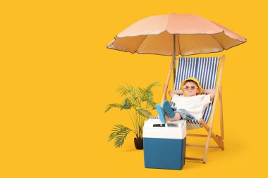 Güverte sandalyesinde oturan sevimli küçük çocuk plaj soğutucusu ve sarı arka planda şemsiyesi var. Seyahat kavramı