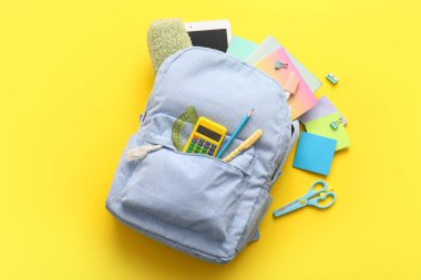 Mavi okul çantasının içinde farklı kırtasiye malzemeleri ve sarı arka planda modern tablet bilgisayar var.