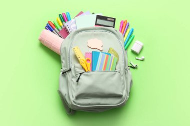 Değişik kırtasiye malzemeleri ve yeşil arka planda modern kulaklıklar olan şık bir okul çantası.