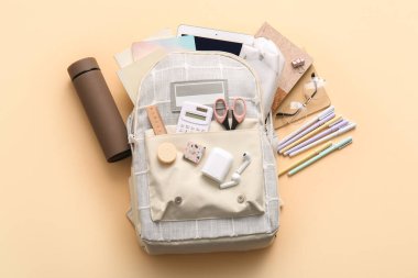 Farklı kırtasiye malzemeleri, termos ve bej arka planda modern tablet bilgisayar içeren şık okul çantaları.