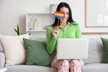 Renkli kağıt kalpli ve modern dizüstü bilgisayarlı güzel bir kadın oturma odasında kanepede oturuyor. LGBT kavramı