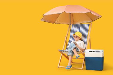 Cep telefonu güverte sandalyesinde oturan ve sarı arka planda plaj soğutucusu olan şirin bir çocuk. Seyahat kavramı