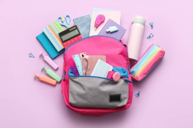 Değişik kırtasiye malzemeleriyle süslü okul çantası ve leylak arka planda su şişesi.