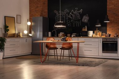 Tezgahları, yemek masası ve geceleri parlayan lambaları olan şık bir mutfak.