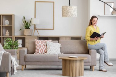 Gülümseyen genç kadın oturma odasındaki gri koltukta dergi okuyor.