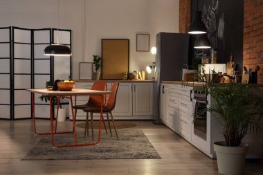 Tezgahları, yemek masası ve geceleri parlayan lambaları olan şık bir mutfak.