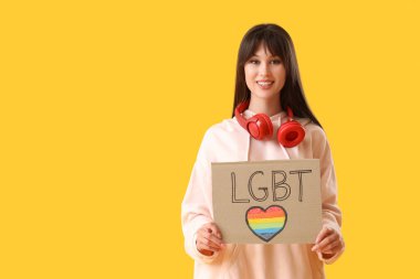 Sarı arka planda LGBT yazan bir pankart tutan güzel bir genç kadın.