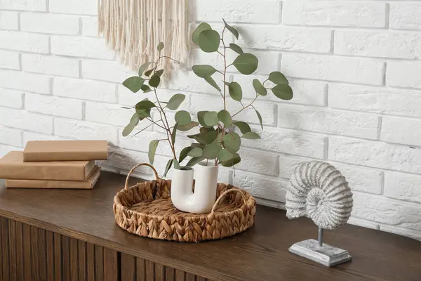 Oturma odasındaki beyaz tuğla duvarın yanındaki masada okaliptüs dalları olan tepsi ve vazo, yakın plan.