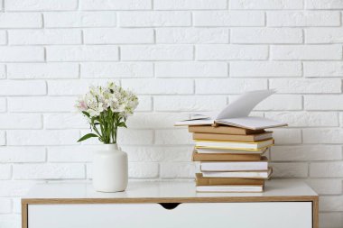 Bir sürü kitap ve vazo. Hafif tuğla duvarın yanındaki çekmecelerde çiçekler var.