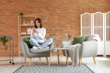 Evdeki yumuşak gri koltukta cep telefonu kullanan genç kadın.