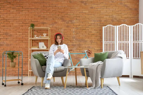 Evdeki gri koltukta cep telefonu kullanan kulaklıklı genç bir kadın.