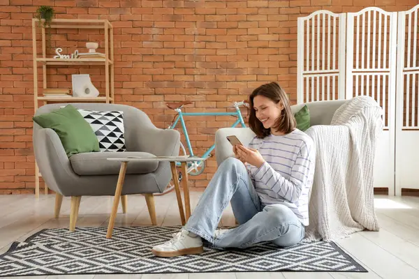 Evdeki koltukların yanındaki halıda cep telefonu kullanan genç bir kadın.