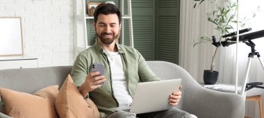 Bilgisayarlı yakışıklı adam evdeki koltukta cep telefonu kullanıyor.