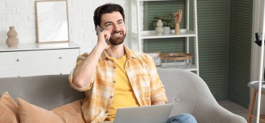 Bilgisayarlı yakışıklı adam evdeki koltukta cep telefonuyla konuşuyor.