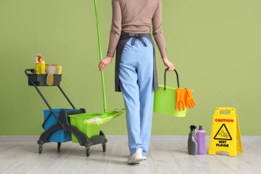 Elinde temizlik malzemeleri ve yeşil duvarın yanındaki uyarı levhası olan kadın hademe.