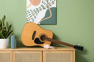 Akustik gitar, kulaklıklar, kitaplar ve ev bitkisi renkli duvarın yanındaki ahşap dolapta.