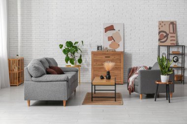 Beyaz tuğla duvarın yanında rahat gri kanepesi, koltuğu ve masası olan bir oturma odası.