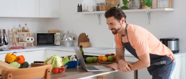 Mutfakta dizüstü bilgisayarı olan yakışıklı sakallı adam.