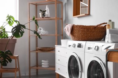 Çamaşır makineleri, hasır sepeti ve temizlik malzemeleriyle çamaşır odasının içinde.