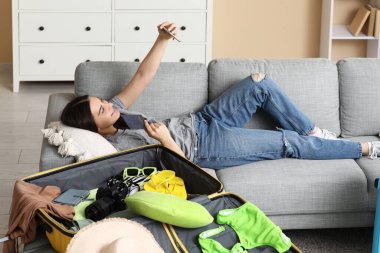 Genç bir kadın cep telefonuyla selfie çekiyor. Evdeki kanepede bavulun yanında. Yaz yolculuğu konsepti