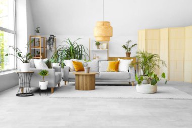 Yeşil bitkilerle, kanepelerle ve masalarla aydınlık oturma odasının içinde.