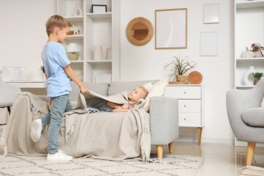 Küçük çocuk evde kız kardeşini ekoseyle örtüyor.