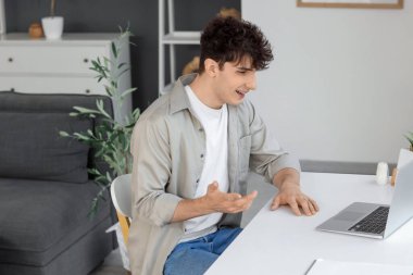Genç adam evdeki masada dizüstü bilgisayarla sohbet ediyor.