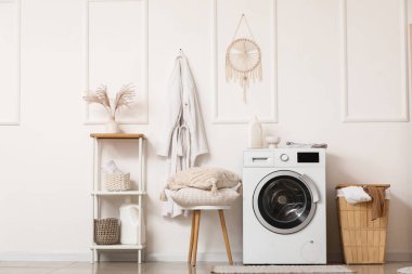 Çamaşır sepetli ve yastıklı modern çamaşır makinesi banyodaki beyaz duvarın yanındaki taburede.