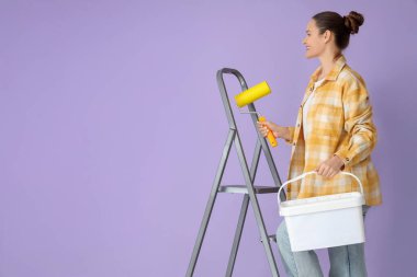 Leylak arka planına karşı merdiven üzerinde boya silindiri ve kova taşıyan genç bir kadın.