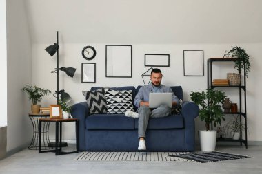 Genç adam mavi rahat koltukta oturuyor ve oturma odasında dizüstü bilgisayarla çalışıyor.