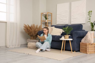 Genç bir kadın kitap okuyor ve oturma odasındaki mavi kanepenin yanında oturuyor.