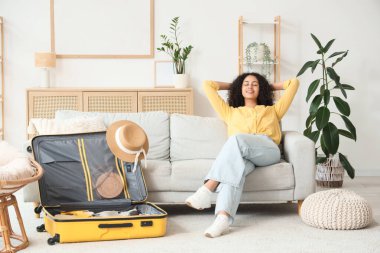 Evdeki kanepede oturan bavullu genç Afro-Amerikalı kadın. Seyahat kavramı