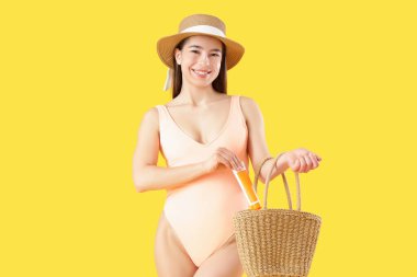 Mayolu genç bir kadın sarı arka planda plaj çantasına güneş kremi sürüyor.