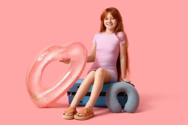 Şişme yüzüklü ve boyun yastıklı şirin bir kız pembe arka planda bavulun üzerinde oturuyor. Seyahat kavramı