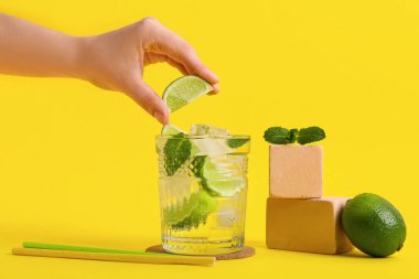 Kadın eli, sarı arka planda taze mojito kokteyli bardağına limon sıkıyor.