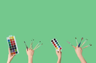 Artistlerin fırçaları, palet bıçakları ve yeşil arka planda suluboya boyalı kadın elleri.