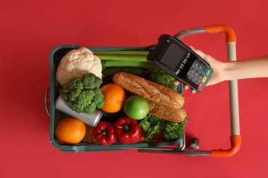 Kırmızı arka planda ödeme terminali olan yiyecek ve kadın eli dolu bir alışveriş arabası.