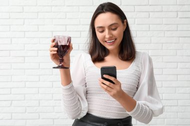 Beyaz tuğlalı arka planda kırmızı şarap ve cep telefonuyla güzel bir genç kadın.