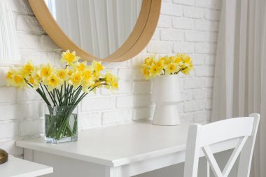 Beyaz odadaki masada sarı narsisli çiçekler olan vazolar.