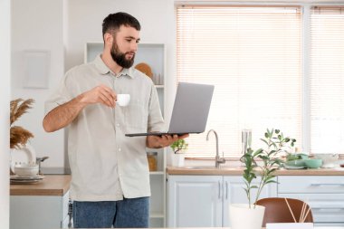 Kahve fincanı ve dizüstü bilgisayarlı genç sakallı adam mutfakta çalışıyor.