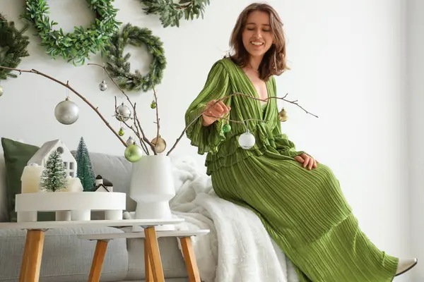 Oturma odasındaki ağaç dallarında Noel topları olan genç bir kadın.