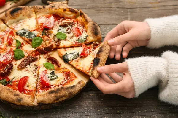 Ahşap arka planda lezzetli pizzadan bir dilim alan kadının elleri, yakın plan.
