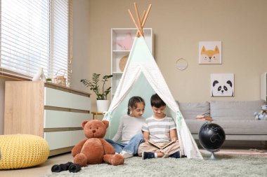 Evdeki çadırda macera kitabı okuyan sevimli çocuklar.