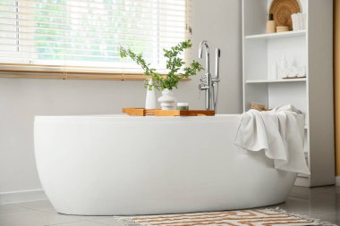 Küveti, raf ünitesi ve pencerenin yanındaki vazoda çiçek açan yeşil dalları olan şık bir banyo.