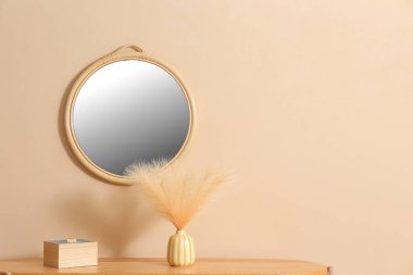 Kutu, vazo, dekoratif tüyler ve bej duvarda yuvarlak ayna, yakın plan.