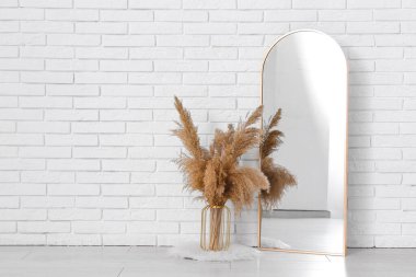 Odanın içinde beyaz tuğla duvarın yanında otlu vazo ve ayna.